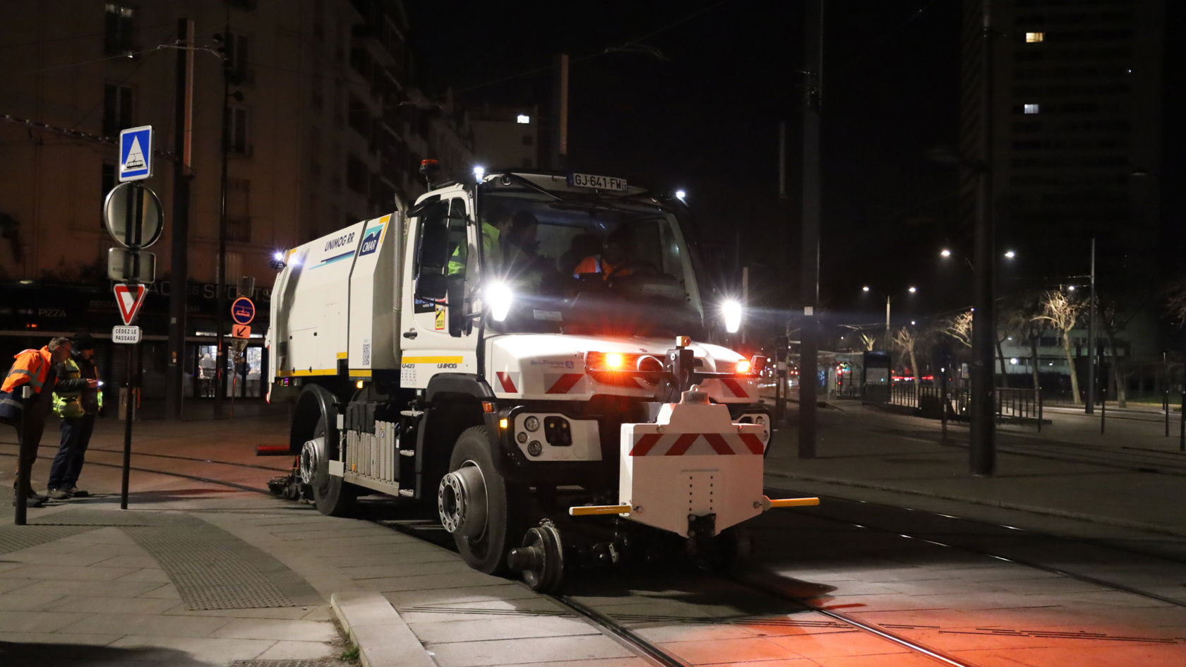 白色 Unimog 带铁路装备，夜间在城市环境中的电车路段上使用。